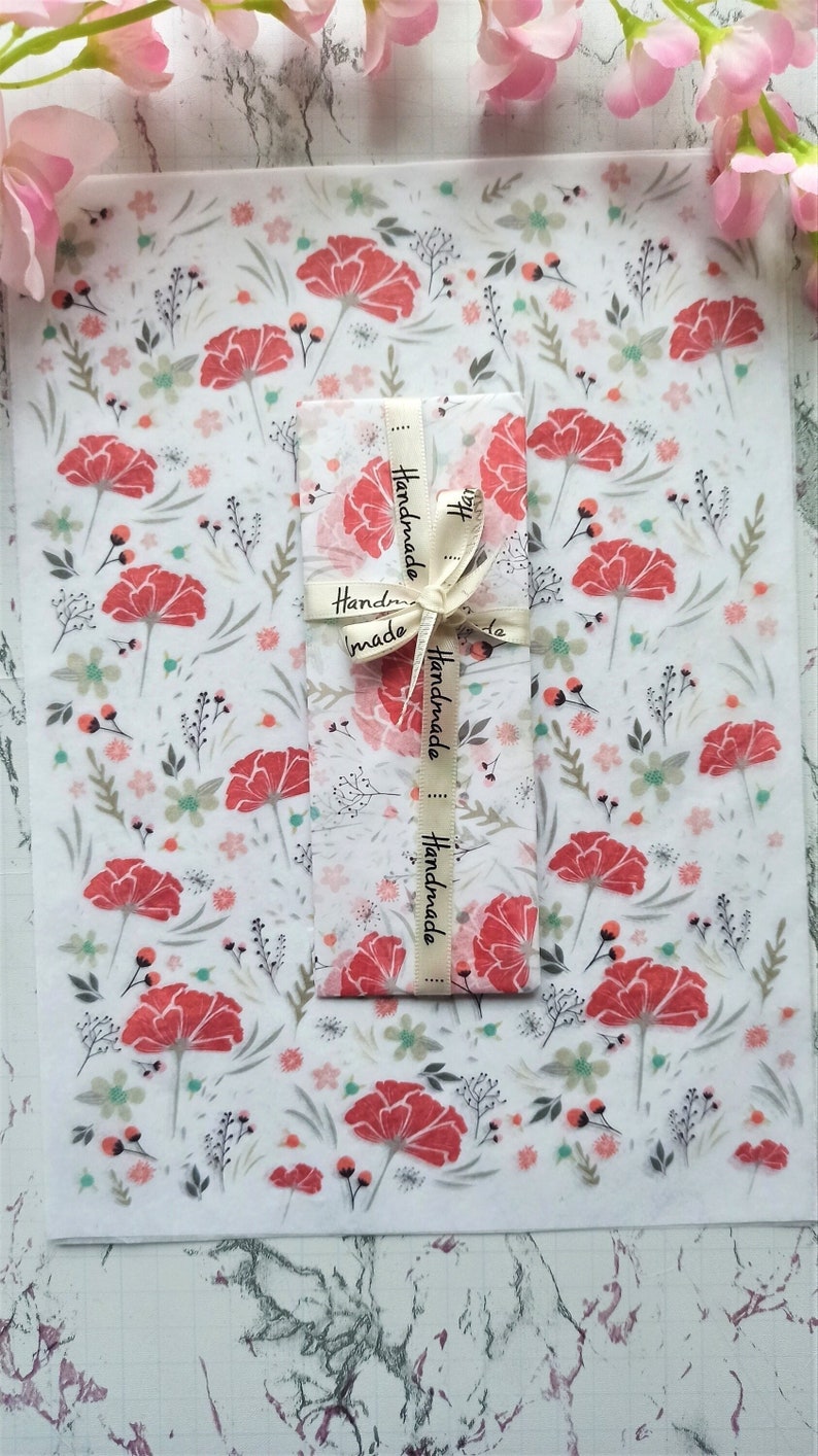 May include: A white sheet of paper with a floral pattern featuring red flowers and green leaves. The paper is wrapped around a smaller sheet of paper with a white ribbon that says "Handmade" four times.
