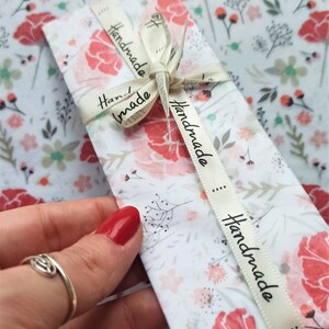 May include: A white envelope with a floral pattern and a cream ribbon tied around it. The ribbon has the word "Handmade" printed on it in black.
