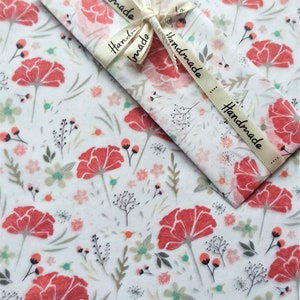 May include: White tissue paper with a floral pattern featuring red flowers and green leaves. The tissue paper is wrapped around a gift with a white ribbon that says "Handmade" in black lettering.
