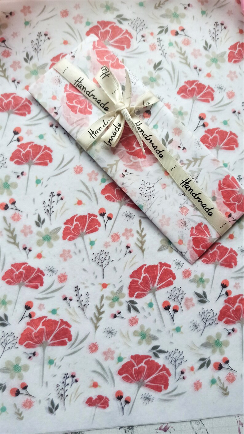 May include: White tissue paper with a floral pattern featuring red flowers and green leaves. The tissue paper is wrapped around a gift with a white ribbon that says "Handmade" in black lettering.