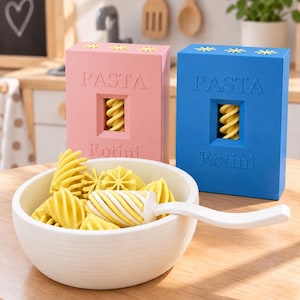 Könnte beinhalten: Eine weiße Schüssel gefüllt mit gelben Rotini-Nudeln und einem weißen Servierlöffel, begleitet von zwei rechteckigen Schachteln in Rosa und Blau, jeweils beschriftet mit PASTA und Rotini, mit einem Sichtfenster für ein Rotini-Stück.