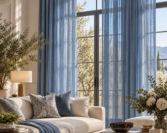 May include: Floor-to-ceiling blue sheer curtains hang in front of a large window. A beige sofa sits in front of the curtains, featuring a blue patterned throw pillow, a solid blue pillow, and a blue throw blanket draped over the seat.