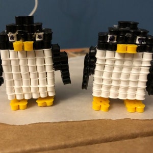 Puede incluir: Dos pequeñas figuras de pingüinos hechas de cuentas blancas y negras con picos y patas amarillas. Los pingüinos están de pie sobre una superficie blanca.
