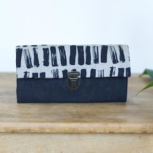 Op de afbeelding: Een rechthoekige, marineblauwe clutch met een klep met een zwart-wit penseelstrekenpatroon. De clutch heeft een metalen sluiting en ligt op een houten oppervlak. Het thema zijn accessoires.