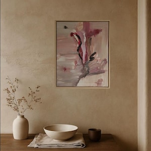 Op de afbeelding: Een abstract schilderij in een gouden lijst, met tinten roze, rood en zwart. Een witte vaas met gedroogde bloemen, een witte kom en een kleine bruine beker staan op een houten tafel. De achtergrond is neutraal beige.