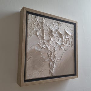 Könnte beinhalten: Quadratisches abstraktes Kunstwerk mit strukturierter, erhabener Oberfläche. Das Kunstwerk zeigt eine neutrale Farbpalette aus Beige und Weiß, gerahmt in einem Naturholzrahmen mit schwarzem Innenrand. Das Kunstwerk ist an einer weißen Wand befestigt.