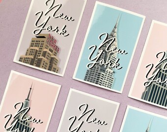 Puede incluir: Seis postales con diferentes imágenes de los rascacielos de la ciudad de Nueva York. Cada postal tiene un fondo rosa, azul o morado claro con el texto "Nueva York" escrito en cursiva negra.