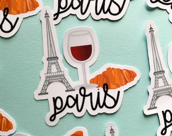 Puede incluir: Un conjunto de pegatinas con temática parisina que incluyen la Torre Eiffel, croissants, copas de vino y la palabra "paris" en una fuente manuscrita. Las pegatinas están sobre un fondo azul claro.