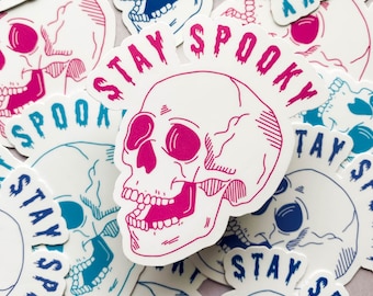 Puede incluir: Una colección de pegatinas con una calavera rosa y azul con el texto "Stay Spooky" en una fuente que gotea. Las pegatinas están sobre un fondo blanco.