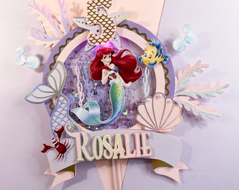 May include: A purple and pink cake topper featuring Ariel from The Little Mermaid, a seashell, a starfish, and a crab. The topper also includes a banner with the name "ROSALIE" and a number "5" with a mermaid tail design.
