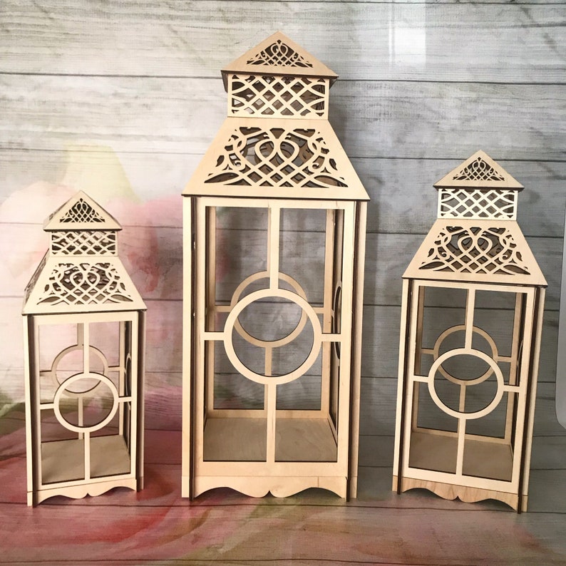 May include: Three wooden candle lanterns of varying sizes with intricate cut-out designs. The lanterns are made of natural wood and have a rustic, farmhouse style.
