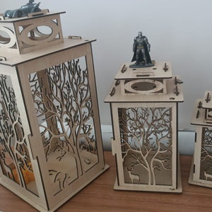 May include: Three wooden candle lanterns with intricate tree designs. The lanterns are made of light brown wood and have a square shape. The largest lantern is about 12 inches tall and the smallest is about 6 inches tall.