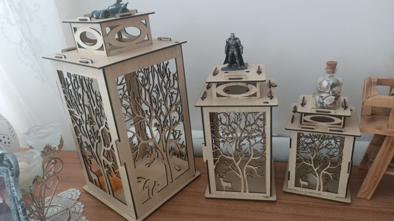 May include: Three wooden candle lanterns with intricate tree designs. The lanterns are made of light brown wood and have a square shape. The largest lantern is about 12 inches tall and the smallest is about 6 inches tall.
