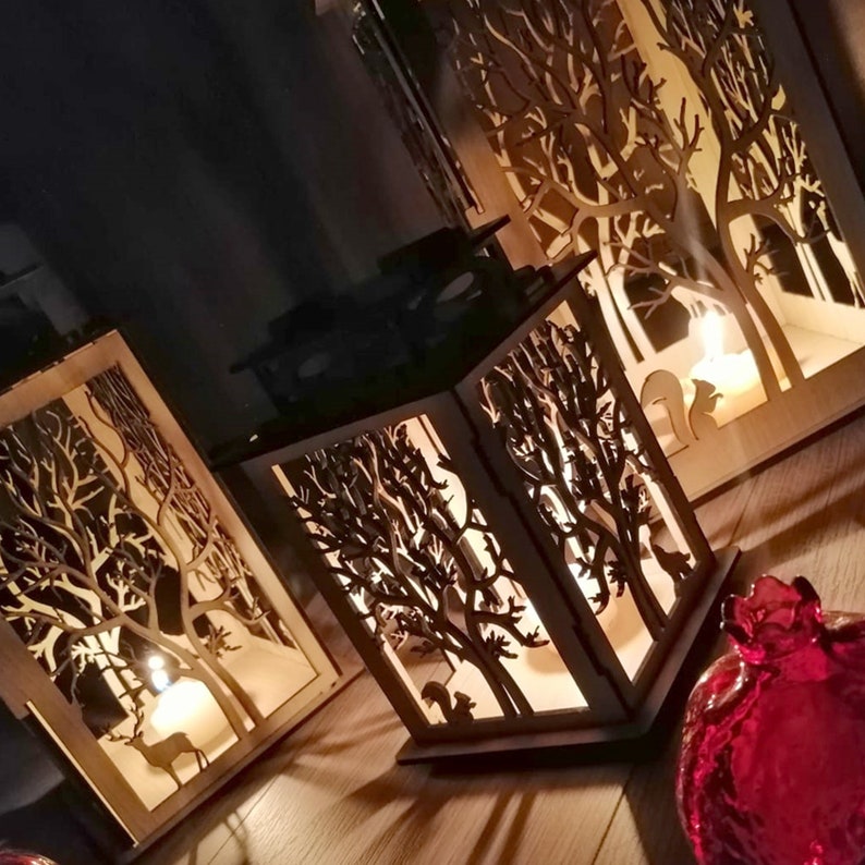 May include: Three wooden candle lanterns with intricate cut-out designs of trees and animals. The lanterns are lit with candles, casting a warm glow.