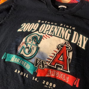May include: A navy blue t-shirt with a baseball graphic. The graphic features the Seattle Mariners logo and the Los Angeles Angels logo. The text "Safeco Field 2009 Opening Day Mariners vs Angels April 14, 2009" is printed on the shirt.