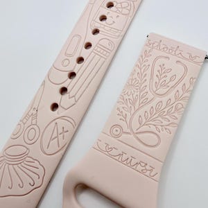 May include: A light pink silicone watch band featuring engraved line art designs. The long strap displays a pencil, scissors, an A+ grade, and a jar. The shorter strap features a stethoscope intertwined with floral elements and the word nurse.