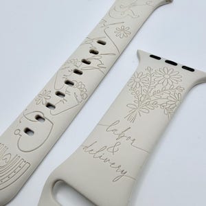 May include: A light beige silicone watch band featuring engraved line art designs. One strap displays a fetus, flowers, and a rainbow. The other strap features a bouquet of daisies and the cursive text labor & delivery, with black metal connectors.