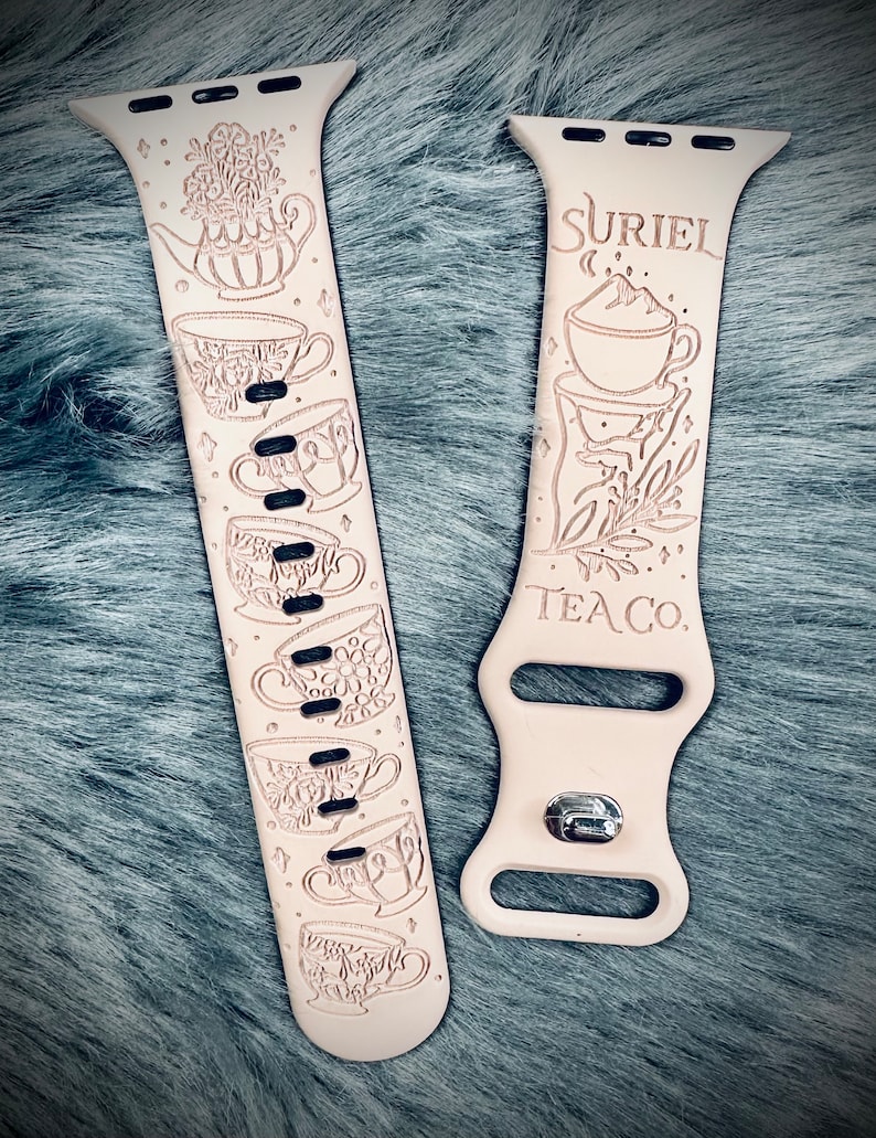 May include: A light beige silicone watch band featuring laser-engraved tea-themed illustrations. One strap displays a teapot and multiple floral teacups, while the other features a teacup, foliage, and the text SURIEL TEA CO. with a metal clasp.
