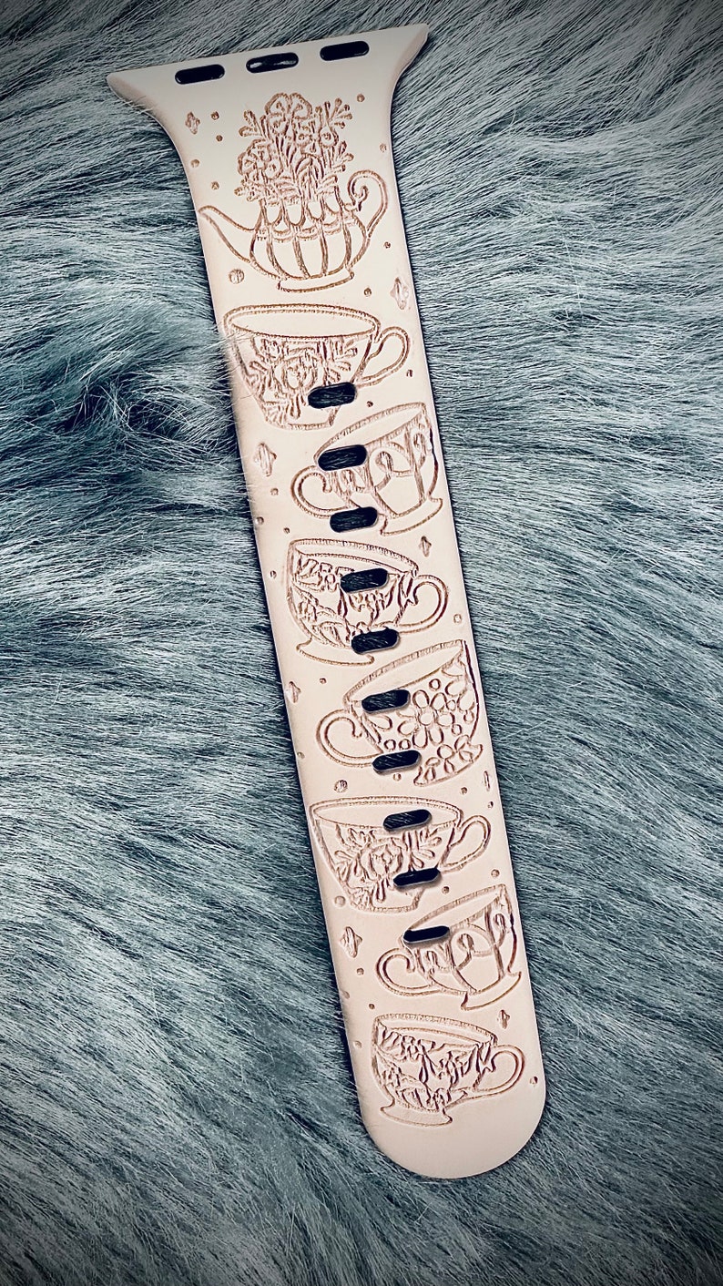 May include: A light beige silicone watch band featuring a laser-engraved pattern of a teapot filled with flowers at the top, followed by a series of six distinct teacups arranged vertically along the length of the strap, interspersed with small star icons.