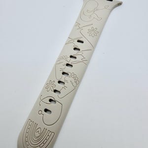 May include: A beige silicone watch band featuring a continuous line art design. The engravings include a baby silhouette, the word baby, two small flowers, a pair of baby footprints, a heart, and a rainbow at the bottom end of the strap.