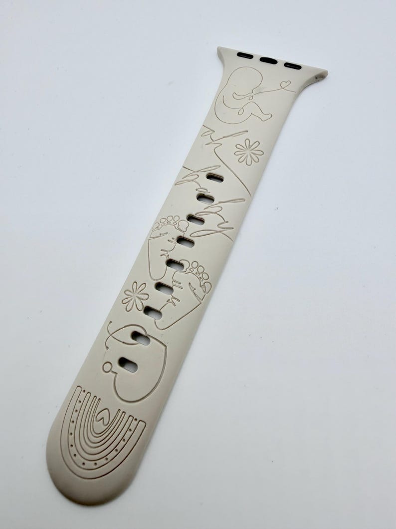 May include: A beige silicone watch band featuring a continuous line art design. The engravings include a baby silhouette, the word baby, two small flowers, a pair of baby footprints, a heart, and a rainbow at the bottom end of the strap.