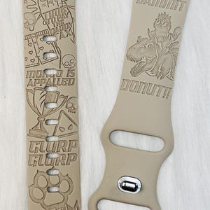 May include: Tan Apple Watch bands with engraved designs and text. One band says "NEW ACHIEVEMENT" and features a hand, skull, and other graphics. The other band says "GOD DAMMIT DONUT!!" with a dinosaur graphic.