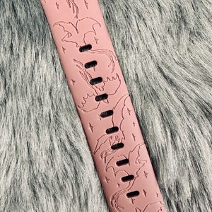 May include: A dusty rose silicone watch band featuring an engraved pattern of multiple flying dragons and small four-pointed stars. The band includes a series of rectangular adjustment holes running down the center and a metal connector at the top.