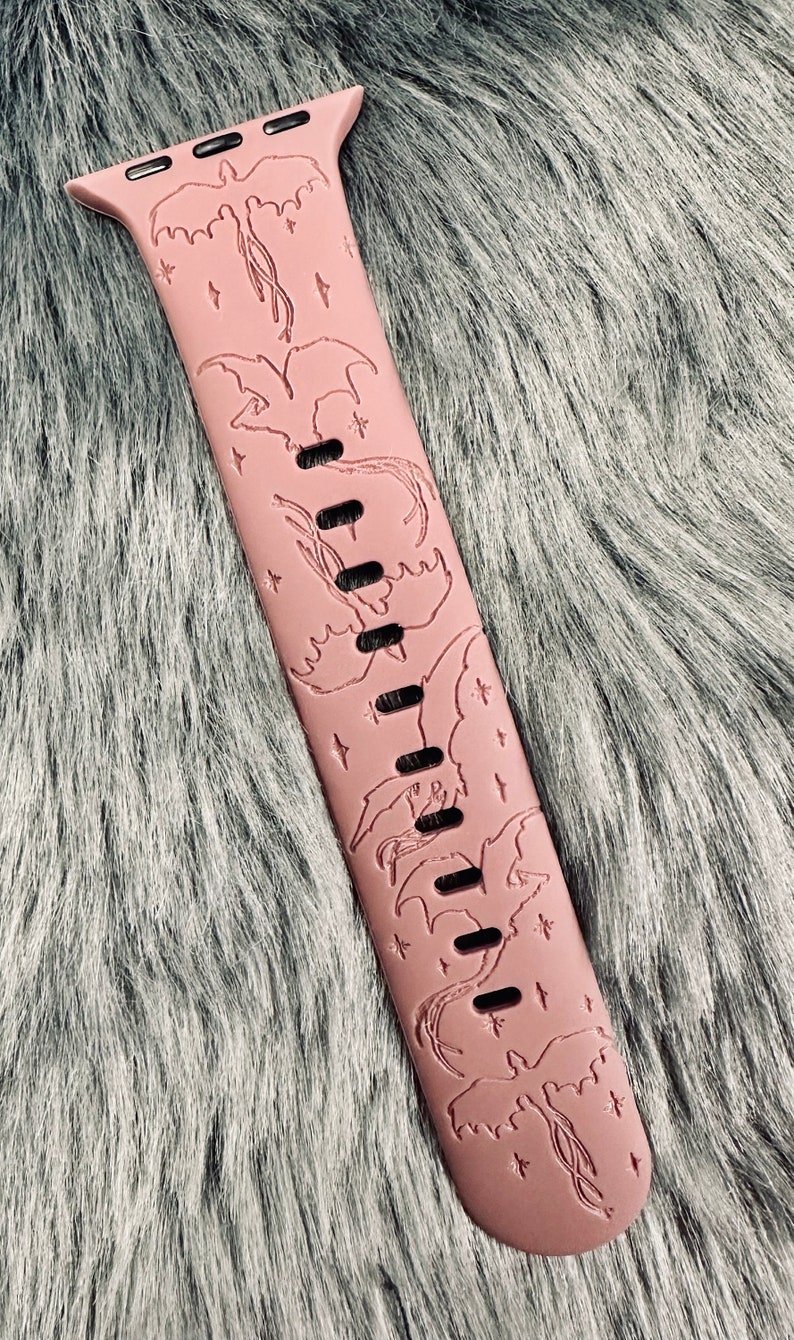 May include: A dusty rose silicone watch band featuring an engraved pattern of multiple flying dragons and small four-pointed stars. The band includes a series of rectangular adjustment holes running down the center and a metal connector at the top.