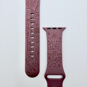 May include: A mauve silicone watch band featuring engraved occult-themed illustrations including a raven, skull, hourglass, and crystals. The text SALEM 1692 THEY MISSED ONE is engraved on the shorter strap along with an eye symbol and a hand.