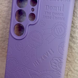 May include: A lavender phone case with embossed text and graphics. The text includes "Princess Donut," "The Queen Anne Chonk," and "III ZEV!!" along with donut and ribbon designs. The case has cutouts for the phone's camera and buttons.