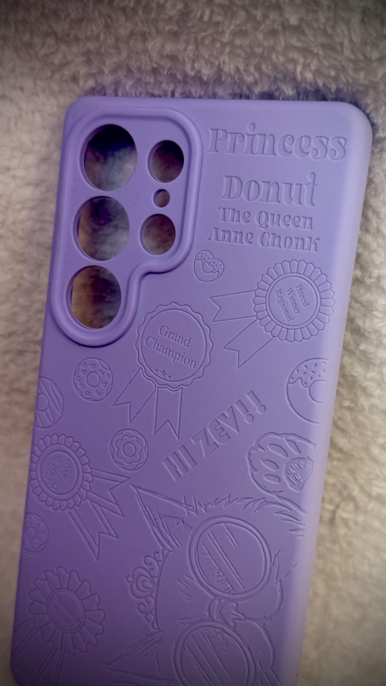 May include: A lavender phone case with embossed text and graphics. The text includes "Princess Donut," "The Queen Anne Chonk," and "III ZEV!!" along with donut and ribbon designs. The case has cutouts for the phone's camera and buttons.