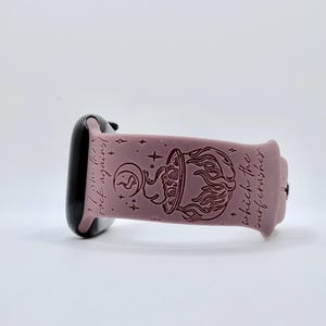 May include: A dusty rose silicone watch band featuring a dark engraved design of a cauldron with flames, a crescent moon, and stars. The text "I am the rock against which the surf crashes" is engraved along the length of the band.