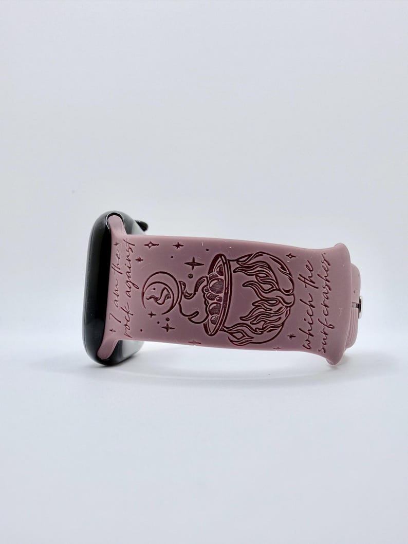 May include: A dusty rose silicone watch band featuring a dark engraved design of a cauldron with flames, a crescent moon, and stars. The text "I am the rock against which the surf crashes" is engraved along the length of the band.