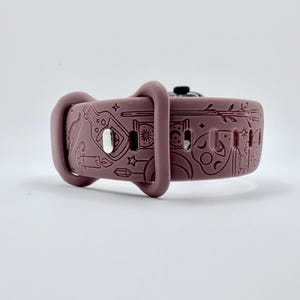 May include: A mauve silicone watch band featuring an embossed pattern of various mystical icons including a potion bottle, a candle, a crescent moon, stars, a wand, and a crystal. The band includes a silver-toned metal pin closure and two loops.