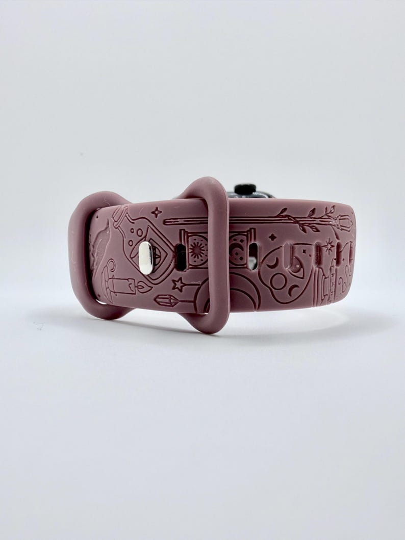 May include: A mauve silicone watch band featuring an embossed pattern of various mystical icons including a potion bottle, a candle, a crescent moon, stars, a wand, and a crystal. The band includes a silver-toned metal pin closure and two loops.