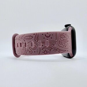 May include: A dusty rose silicone watch band featuring an engraved pattern of celestial and mystical symbols, including a crescent moon, stars, crystals, and a skull. The band is attached to a black smartwatch case with a smooth, matte finish.