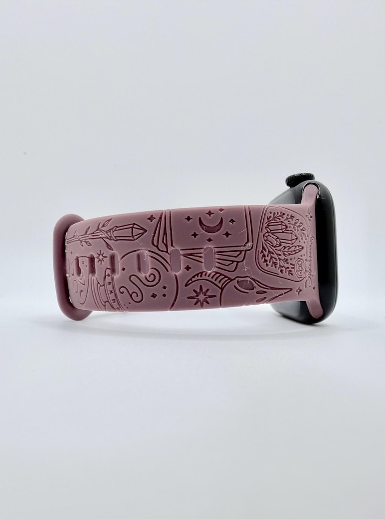 May include: A dusty rose silicone watch band featuring an engraved pattern of celestial and mystical symbols, including a crescent moon, stars, crystals, and a skull. The band is attached to a black smartwatch case with a smooth, matte finish.