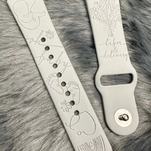 May include: A light gray silicone watch band featuring engraved line art designs including a fetus, baby footprints, flowers, a rainbow, and a stethoscope. The text "oh baby" and "labor & delivery" are engraved on the surface of the two-piece band.