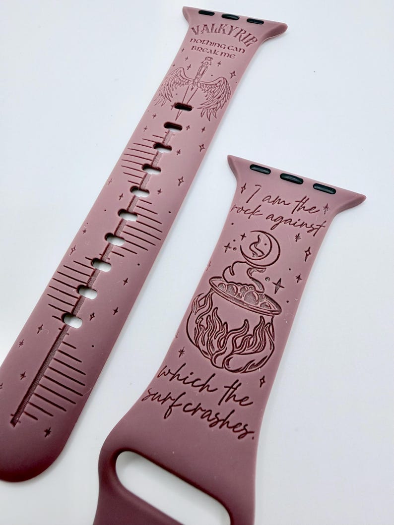 May include: A mauve silicone watch band featuring engraved designs and text. One strap displays a winged sword and the words "VALKYRIE nothing can break me". The other strap shows a cauldron, a crescent moon, and "I am the rock against which the surf crashes".