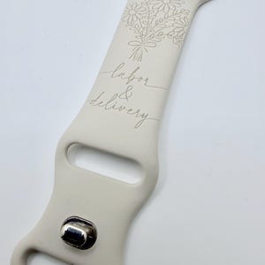 May include: A beige silicone watch band featuring an engraved floral bouquet design at the top and the cursive text labor & delivery centered below. The band includes a metallic silver-toned pin closure and standard attachment hardware at the top.