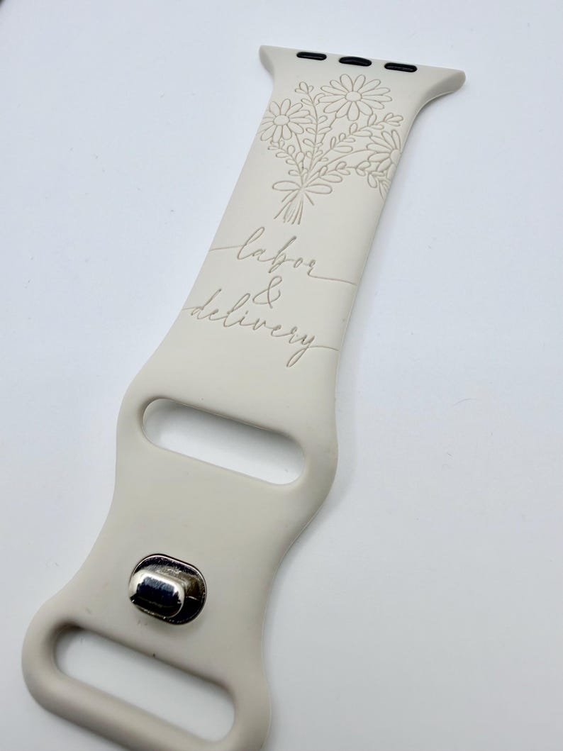 May include: A beige silicone watch band featuring an engraved floral bouquet design at the top and the cursive text labor & delivery centered below. The band includes a metallic silver-toned pin closure and standard attachment hardware at the top.