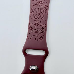 May include: A dark maroon silicone watch band featuring an engraved design of an eye within a triangle, stars, and a hand. The text reads SALEM 1692 THEY MISSED ONE. The band includes a silver-toned metal pin closure and two rectangular slots.