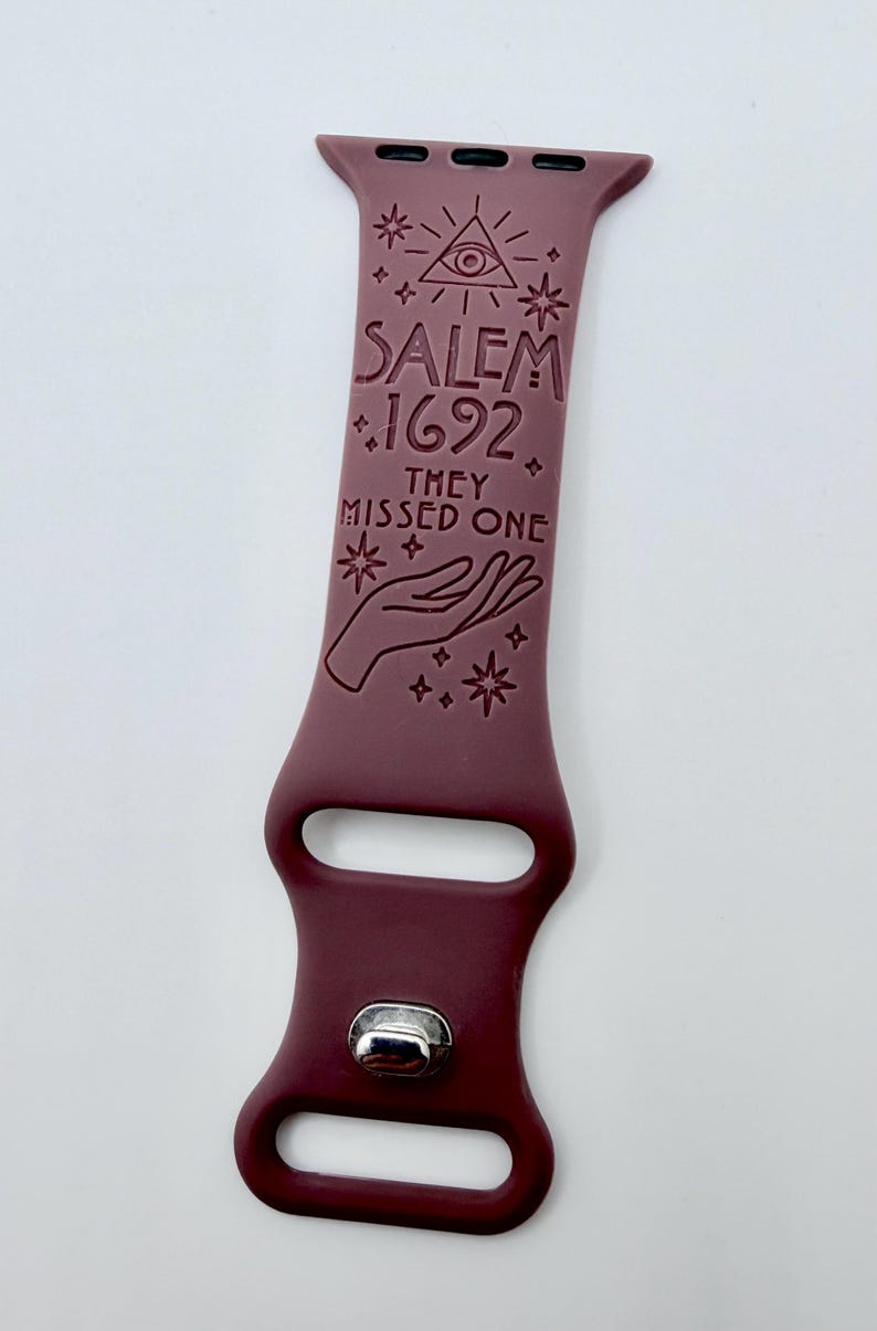 May include: A dark maroon silicone watch band featuring an engraved design of an eye within a triangle, stars, and a hand. The text reads SALEM 1692 THEY MISSED ONE. The band includes a silver-toned metal pin closure and two rectangular slots.