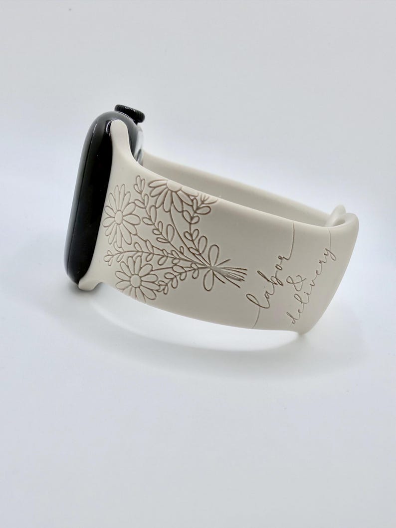 May include: A light beige silicone watch band attached to a black smartwatch case. The band features an engraved floral bouquet design and the cursive text "labor & delivery" etched into the surface, creating a textured, monochromatic appearance.