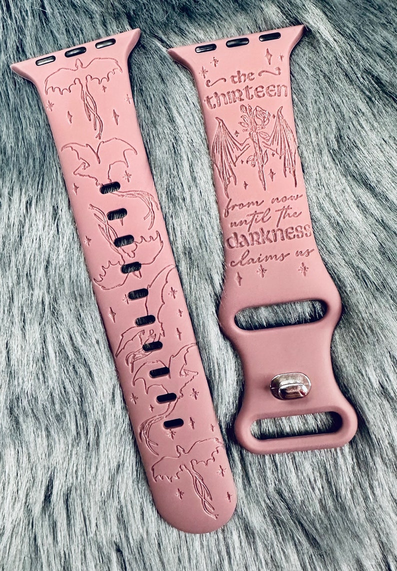 May include: A dusty rose silicone watch band featuring engraved dragon silhouettes and star accents. One side displays the text "the thirteen" above a rose and wing design, with the phrase "from now until the darkness claims us" engraved below.