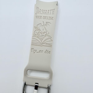 May include: A light beige silicone watch band featuring an engraved design of an open book with a dragon rising from the pages. The text reads Basgiath War College and fly...or die. The band includes a silver-toned metal buckle and a rectangular loop.