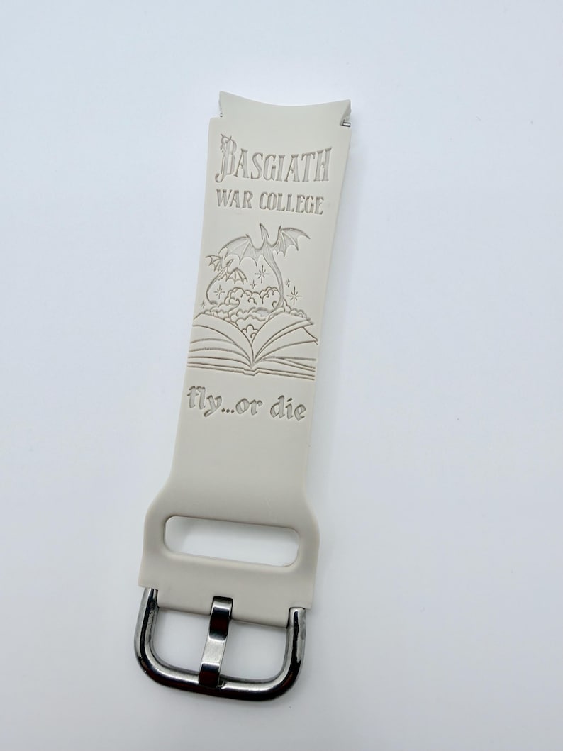 May include: A light beige silicone watch band featuring an engraved design of an open book with a dragon rising from the pages. The text reads Basgiath War College and fly...or die. The band includes a silver-toned metal buckle and a rectangular loop.