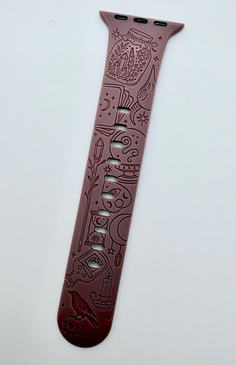 May include: A mauve silicone watch band featuring a laser-engraved pattern of mystical icons including a jar with crystals, a skull, a crystal ball, an hourglass, a potion bottle, a candle, a raven, and various celestial shapes like moons and stars.