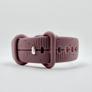 May include: A mauve silicone watch band featuring a debossed pattern of vertical lines and small four-pointed stars. The band includes a matching mauve silicone loop and a polished silver-toned metal pin closure for securing the strap in place.