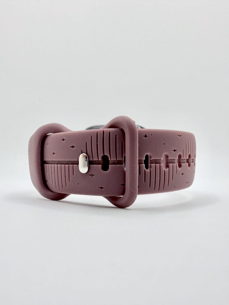 May include: A mauve silicone watch band featuring a debossed pattern of vertical lines and small four-pointed stars. The band includes a matching mauve silicone loop and a polished silver-toned metal pin closure for securing the strap in place.